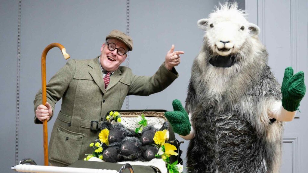 A sheep actor and farmer posing with a pram of newborn lamb plushies at an outdoor event.