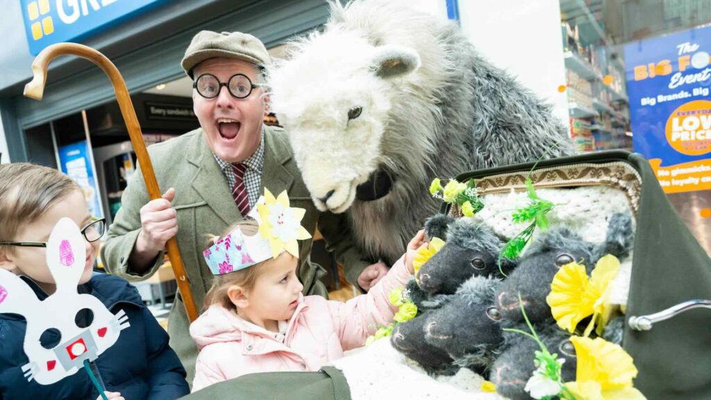 A sheep actor and farmer posing with a pram of newborn lamb plushies and children at an outdoor event.