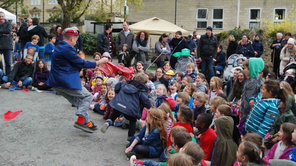 A family circus show at an outdoor public event, with a large crowd of children and families watching the performance.