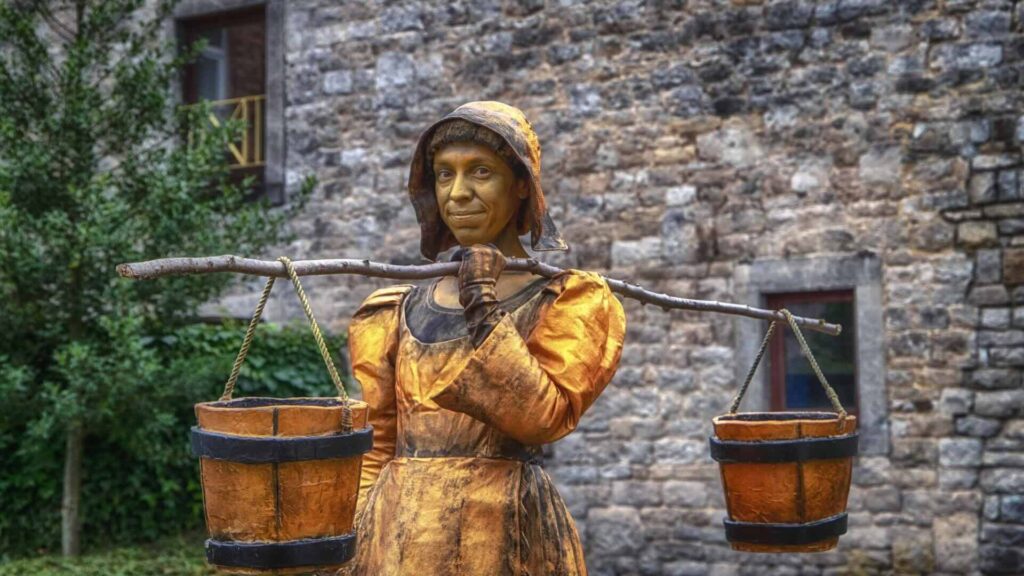 A human metal statue holding two buckets over her shoulder outside of a stoney building.