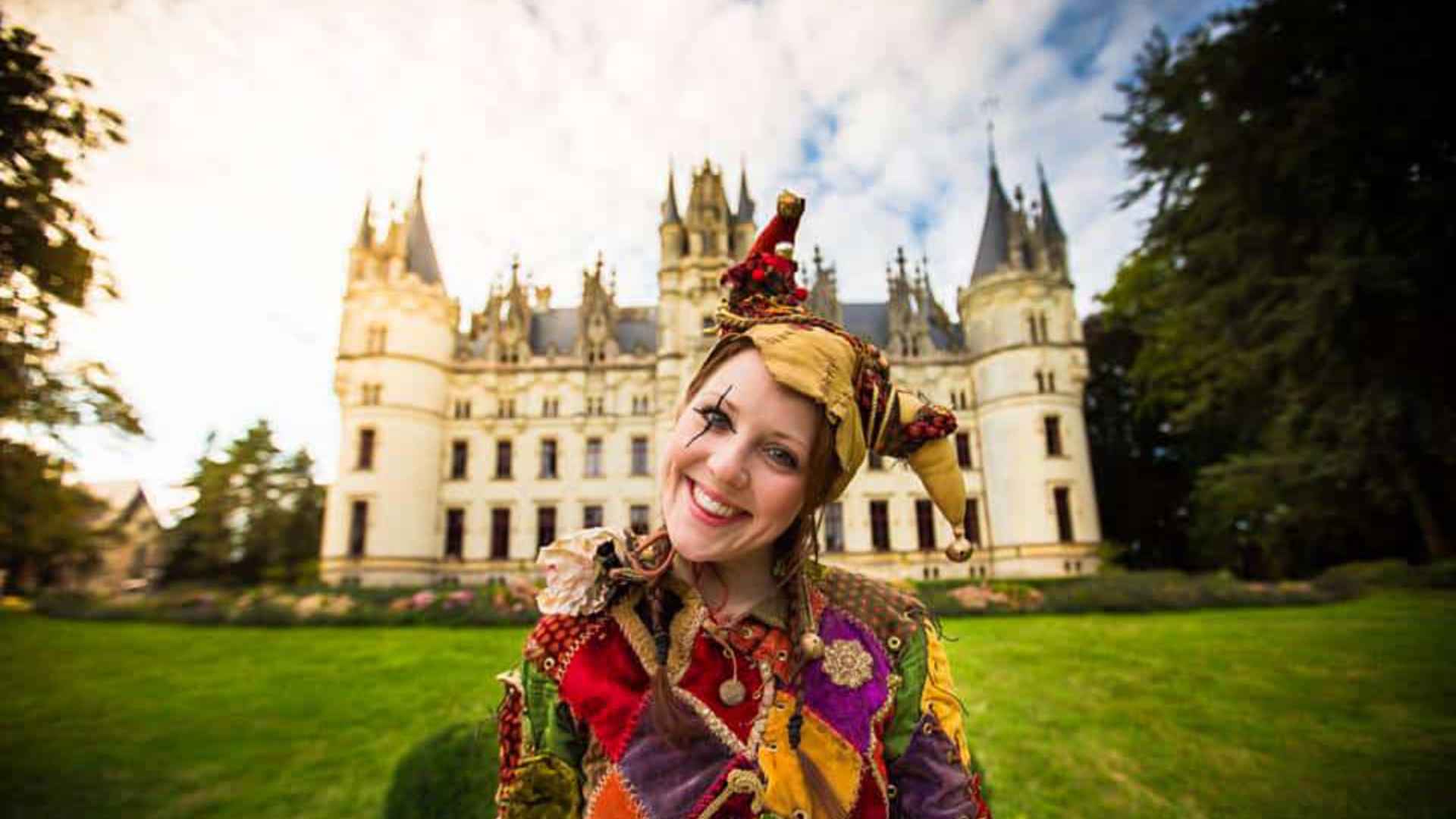 A medieval themed jester stood outside of a castle, looking into the camera.