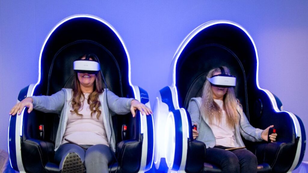 2 women sat in 4d reality pods with vr head set.