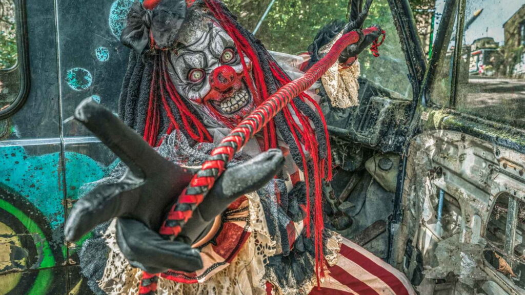 A horror clown performer with red dreadlocks and a striped cane inside a derelict vehicle.