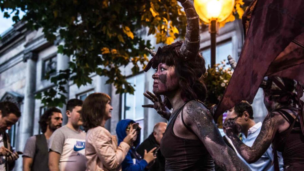 A horned devil performer with wings interacting with a street crowd at an outdoor Halloween event.
