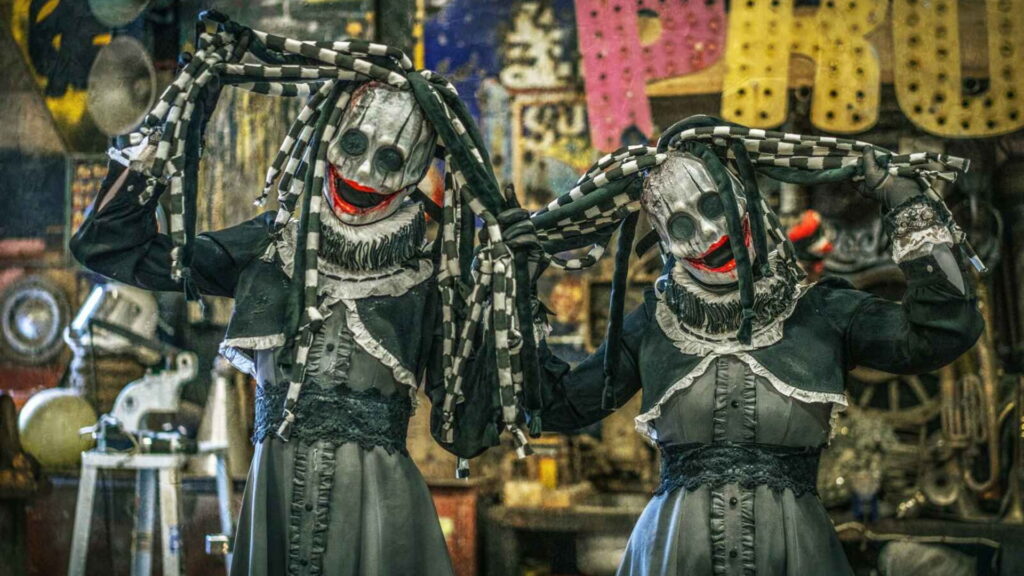 Two identical horror clown performers in monochrome masks and Victorian dress, lifting their chequered dreadlocks.