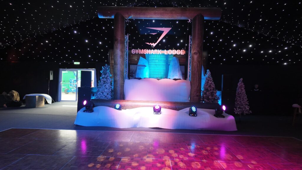Branded Gymshark Lodge stage with timber log portal, snow drift set and moving head lighting at a corporate Christmas party