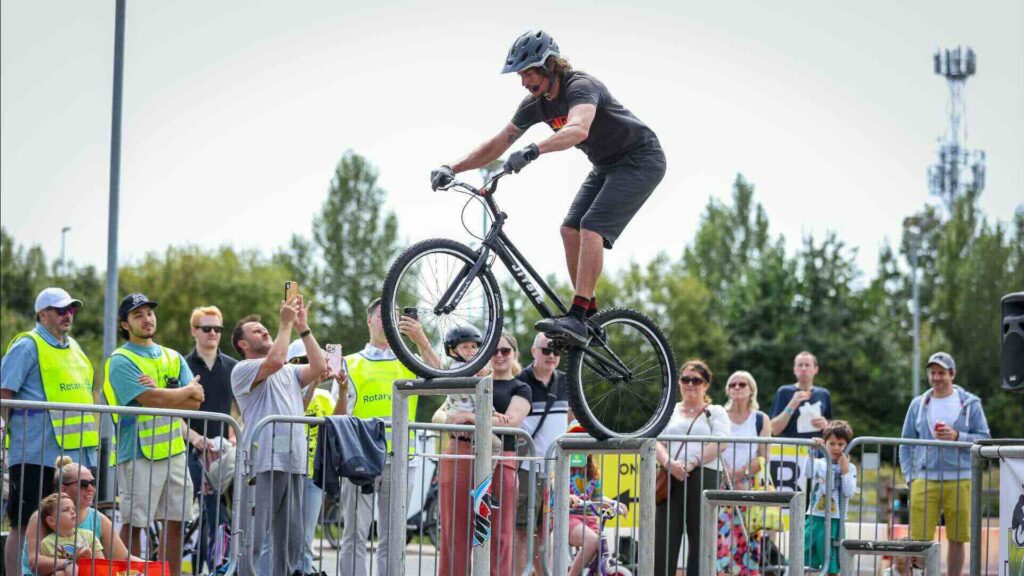 Mountain bike stunt rider balanced on a metal barrier in front of a daytime family crowd at an outdoor event.