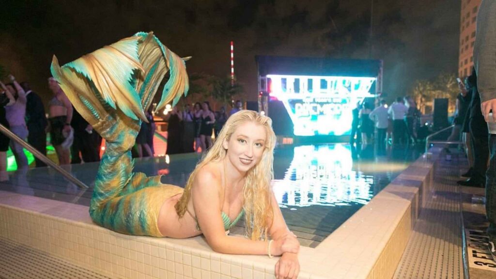 Event mermaid in a green and gold silicone tail posing at the edge of a lit pool during an evening event.