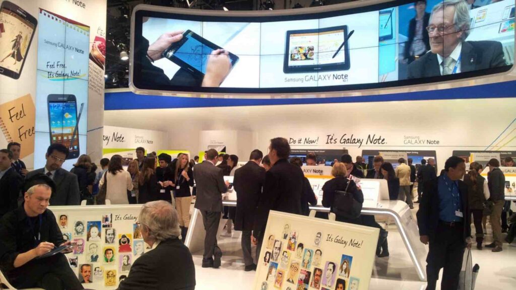 Digital caricaturist drawing on a tablet at a busy exhibition stand with completed caricatures displayed on boards beside him