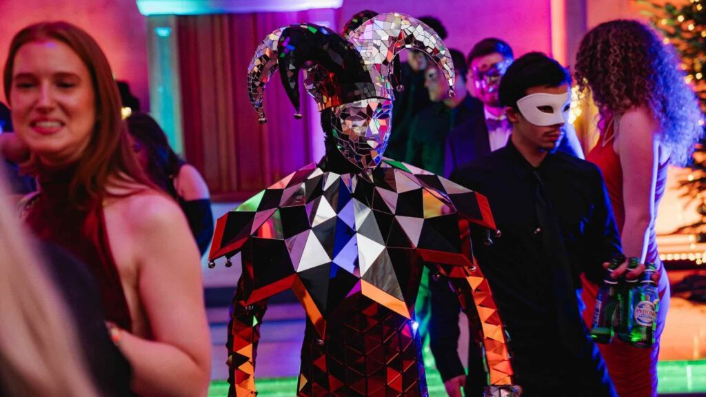Mirror costume walkabout performer in a jester headpiece moving through masked guests at a luxury evening event.