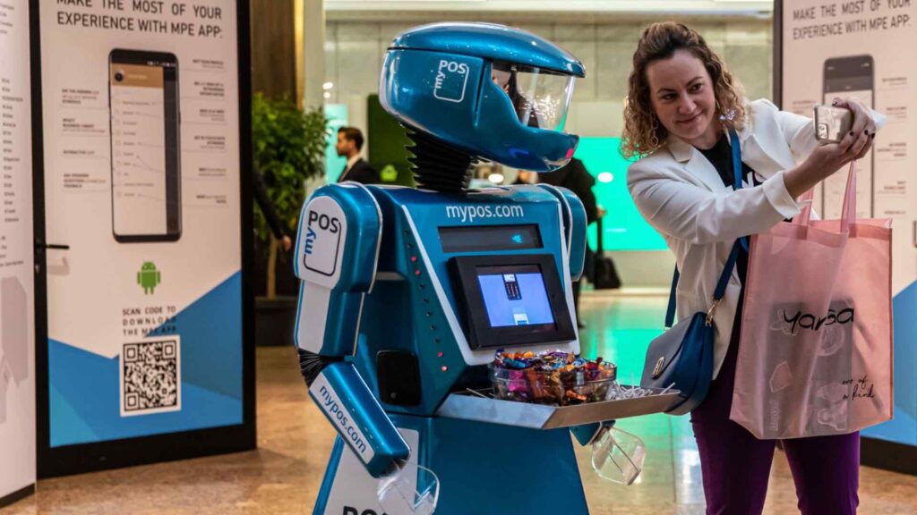 Delegate taking a selfie with a branded promotional event robot holding a tray of giveaways at a convention