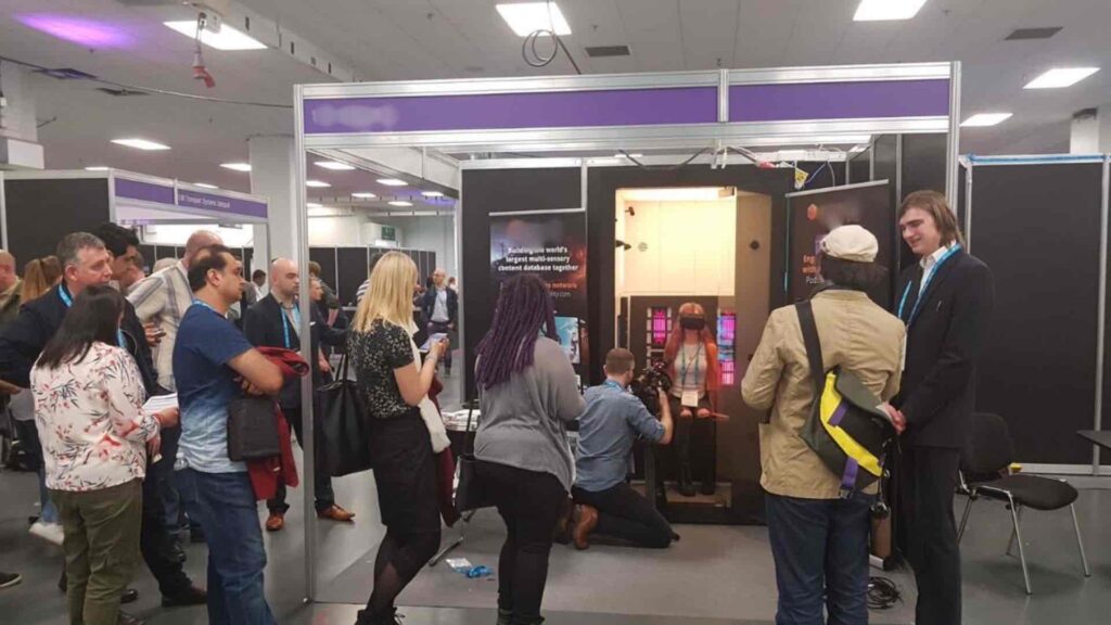 Visitors queuing at an exhibition stand while a delegate wearing a VR headset sits inside an immersive sensory pod