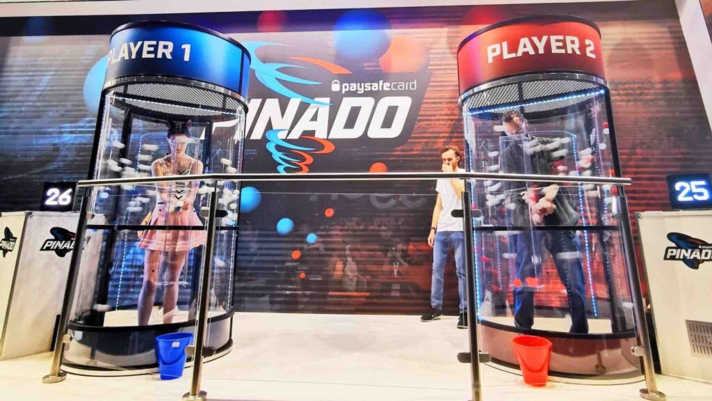 Two players competing inside branded ball grabber pods with a live scoreboard while a visitor watches