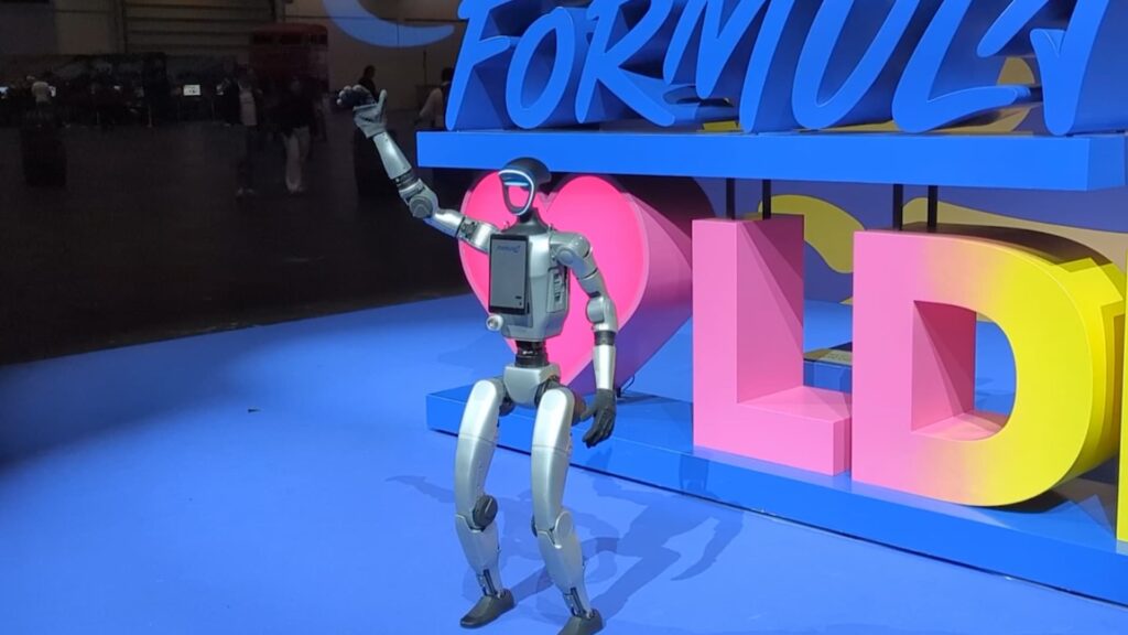 Silver humanoid robot with a light-up visor and chest screen raising one hand to wave, in front of large blue, pink and yellow event lettering
