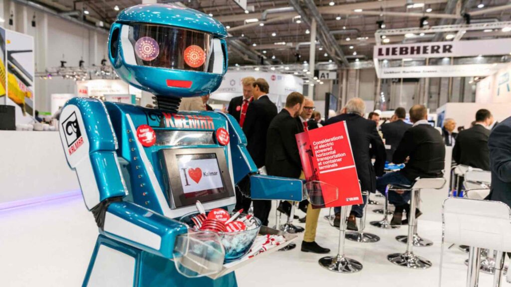 A teal robot with glowing orange eyes standing on an exhibition stand, holding a tray of badges and pens, with a screen on its chest and visitors seated in conversation in the background.