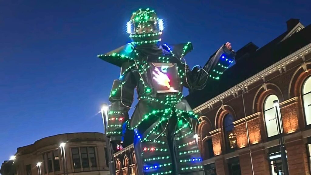 Tall stilt walker in a robot costume covered in green, blue and red LED lights, with a glowing helmet and chest screen, against an evening sky and brick buildings
