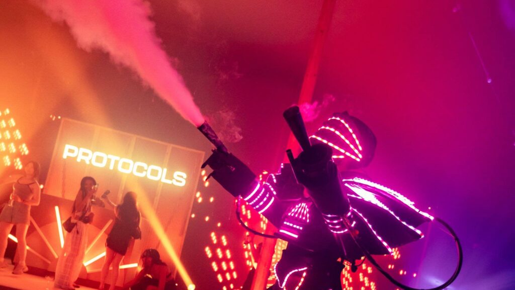 Performer in a robot suit lined with pink LED strips firing a CO2 cannon into the air, with guests and a lit stage in the background