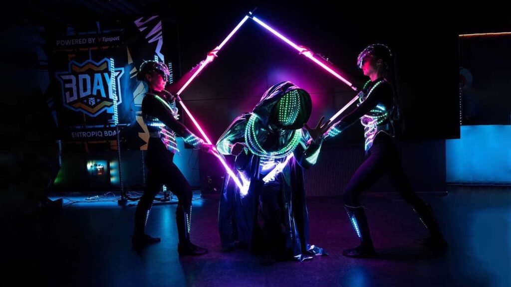 Two performers in LED-lined costumes holding a glowing pink and purple diamond frame around a hooded performer in a light-up mask on a dark stage