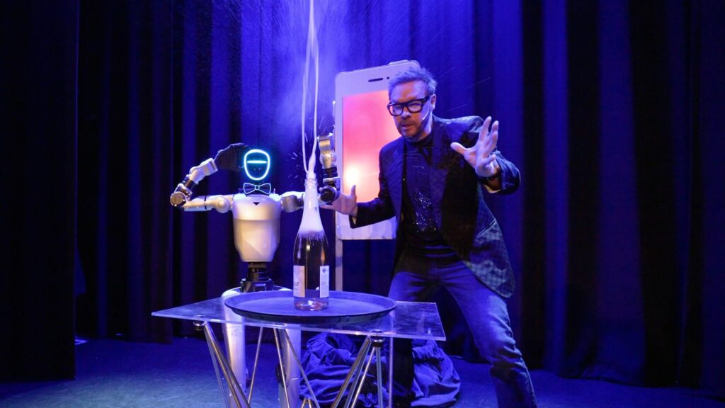 A magician beside a humanoid robot doing their trick as a bottle on a glass table sprays a jet of liquid.