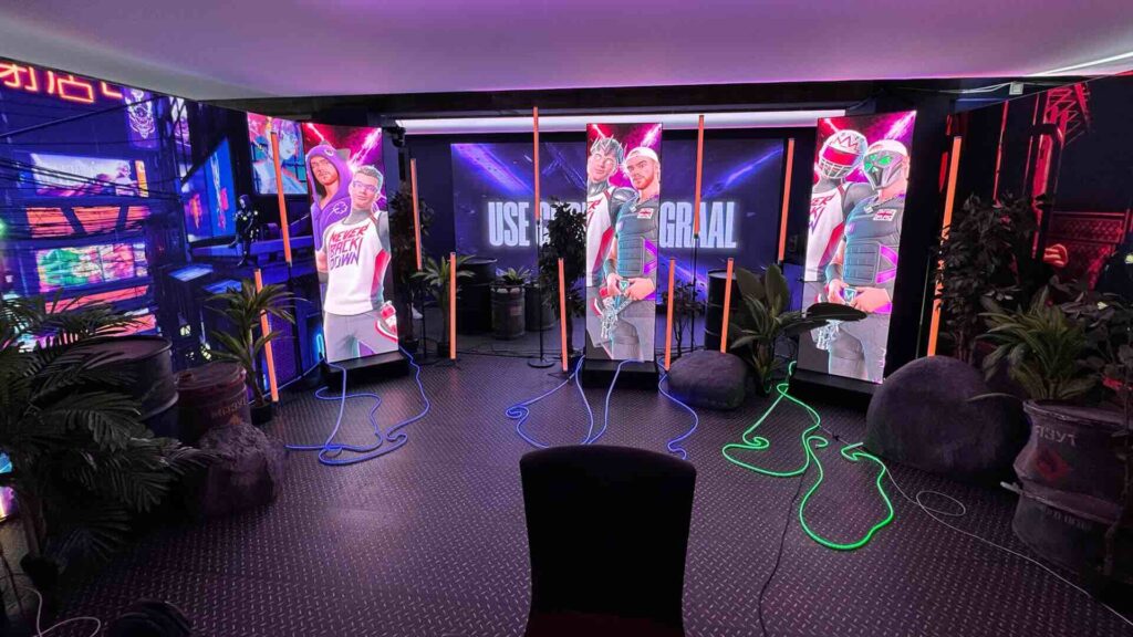 A dark set with three tall portrait LED screens, a large screen behind them displaying a slogan, orange light tubes, and green neon rope lights across a metal floor.