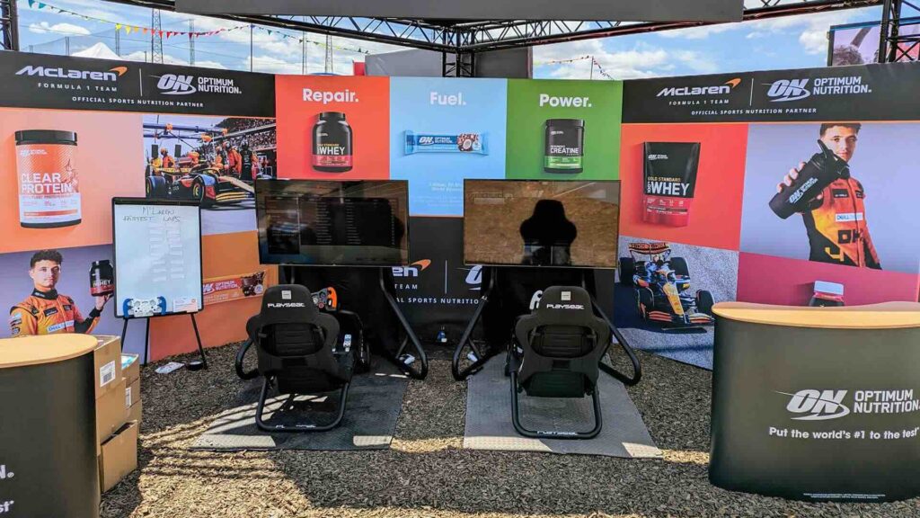 An outdoor exhibition stand with two black racing simulator seats facing screens, colourful product graphics on the back wall.