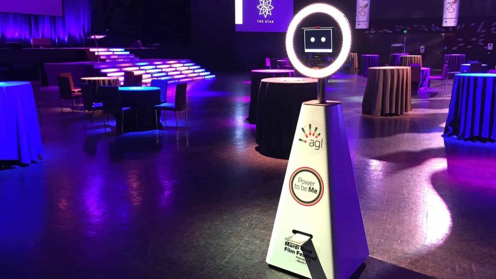 White photo booth robot with a ring light and a screen showing two cartoon eyes, standing in a purple-lit event hall with cocktail tables