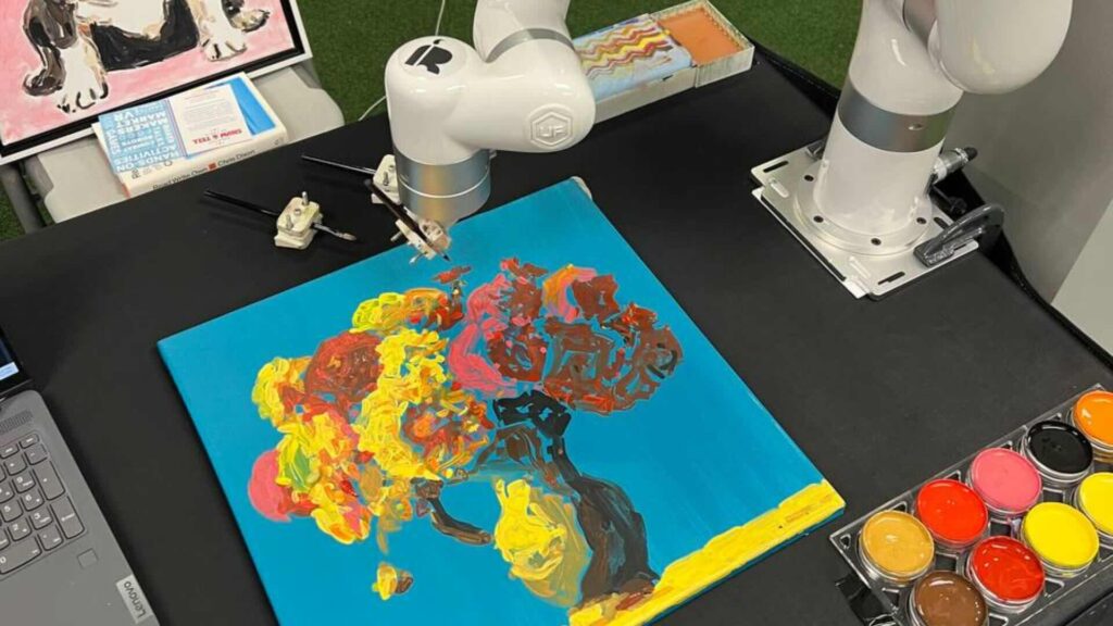 White robotic arm holding a paintbrush over a blue canvas with a colourful painted tree, beside a tray of paint pots