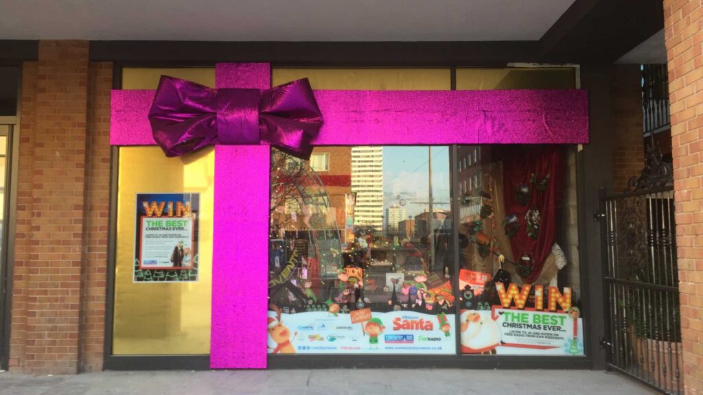 Shop front wrapped like a present with a giant pink glitter ribbon and purple bow above a festive window display