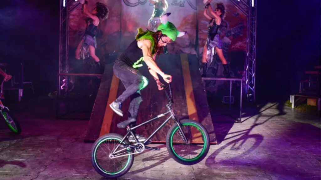 BMX rider in a green helmet balancing on the back wheel on stage, with costumed dancers on platforms behind.