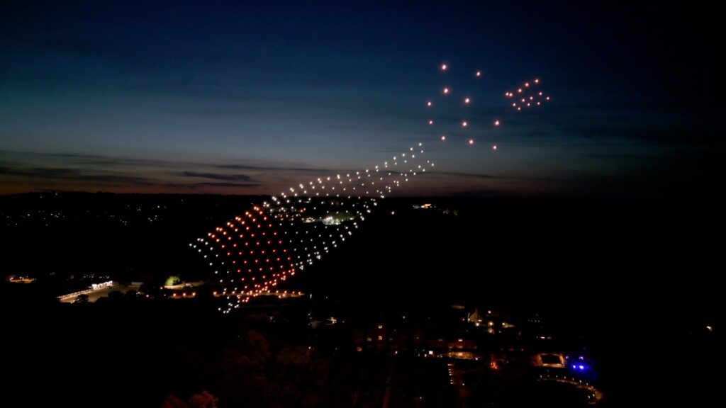 Hundreds of lit drones forming a champagne bottle shape with sparkling bubbles in the dusk sky above a countryside venue