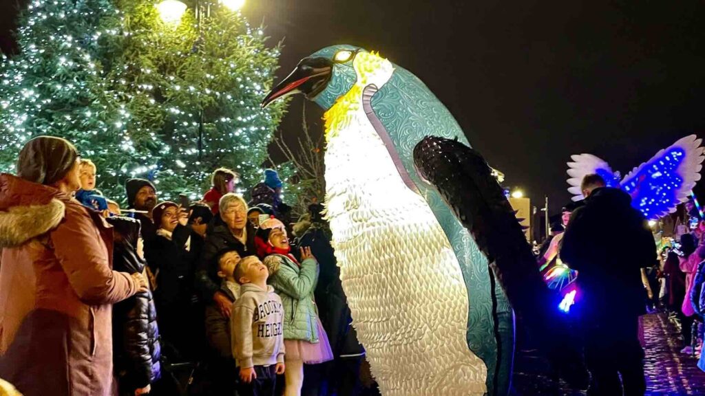 Giant illuminated penguin puppet beside a crowd of families looking up at it, with a lit Christmas tree behind