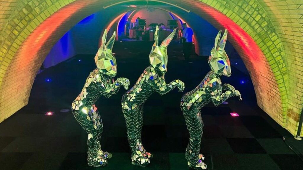 Three performers in mirrored rabbit costumes posing in a brick-arched tunnel lit in red, green and blue