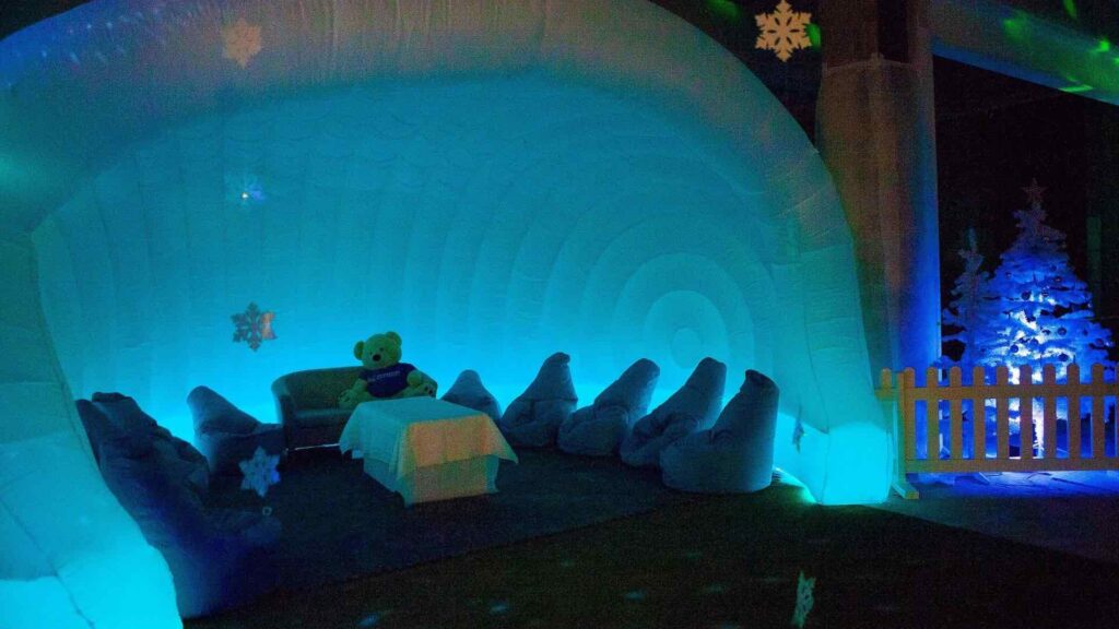Inflatable Christmas dome lit in blue with snowflake projections, bean bag seating and a teddy bear on a sofa
