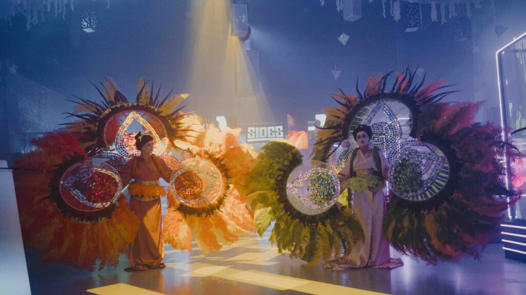 Two performers in silk kimonos holding giant feathered fans decorated with mirror mosaics, under spotlights in a hazy event space